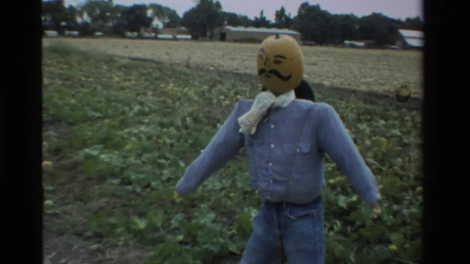 Wider 8mm frame of the pumpkin-headed scarecrow in jeans and a blue shirt, with farm rows and low buildings behind it.