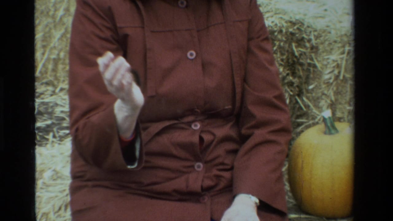 1984 California home-movie frame of a seated woman in a brown coat beside hay and a pumpkin, raising her hand before showing the caterpillar.