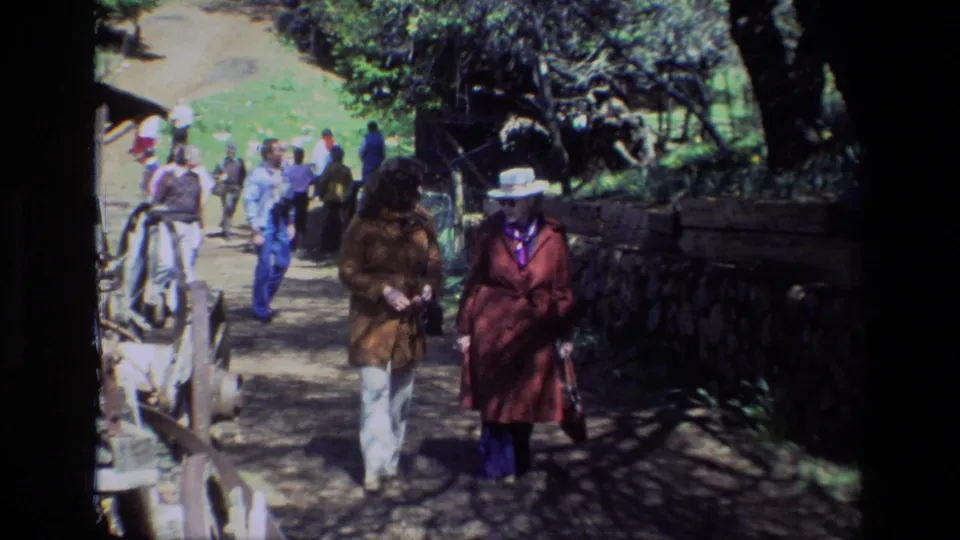 Two visitors in coats approach the camera on a shaded Daffodil Hill trail, with other tourists and rustic landscape features behind them.