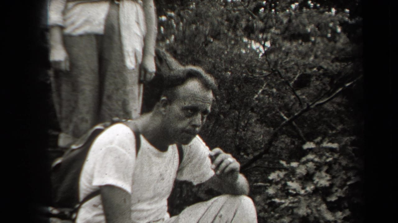 In 1938 New York home-movie footage, a man in a white shirt pauses in dense woodland with a backpack strap over one shoulder.