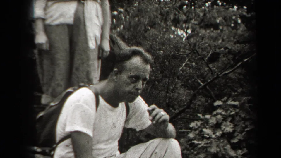 In 1938 New York home-movie footage, a man in a white shirt pauses in dense woodland with a backpack strap over one shoulder.