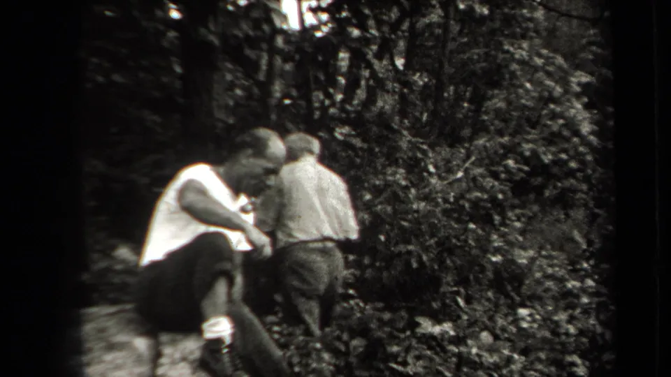 A man in a white shirt sits or crouches near vegetation while another figure moves ahead through the wooded area.