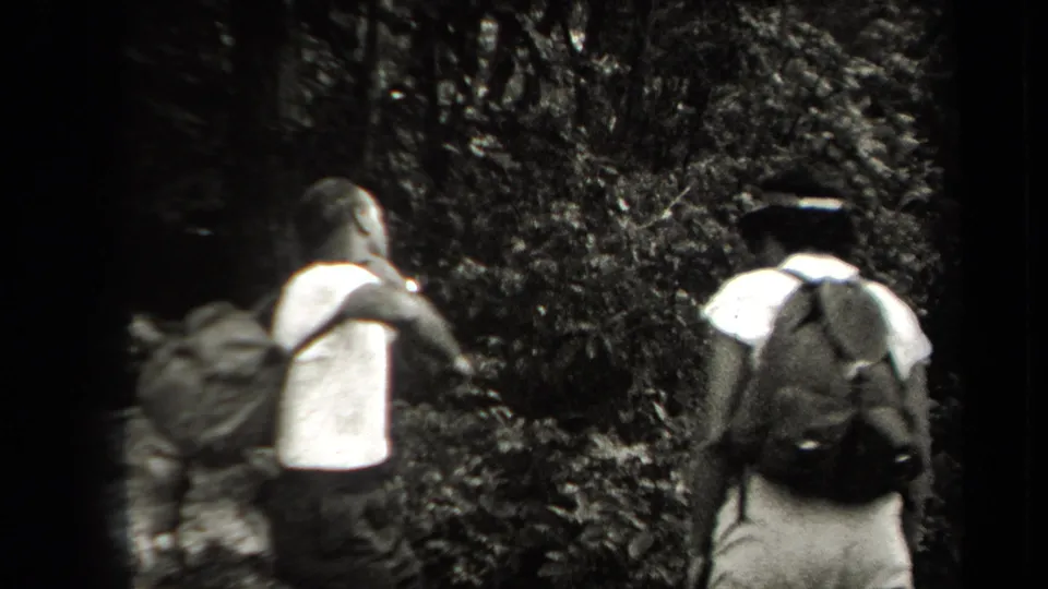 Two adult men with backpacks move away from the camera into dense New York woodland in the 1938 8mm footage.