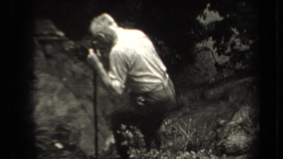 An older adult man in a light shirt bends near rocks and vegetation, holding a walking stick in the wooded setting.