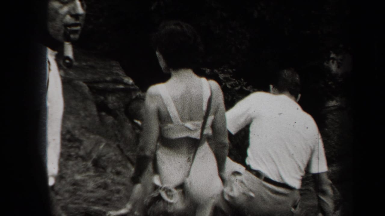 Adults walk near a rocky cave entrance in black-and-white home-movie footage.