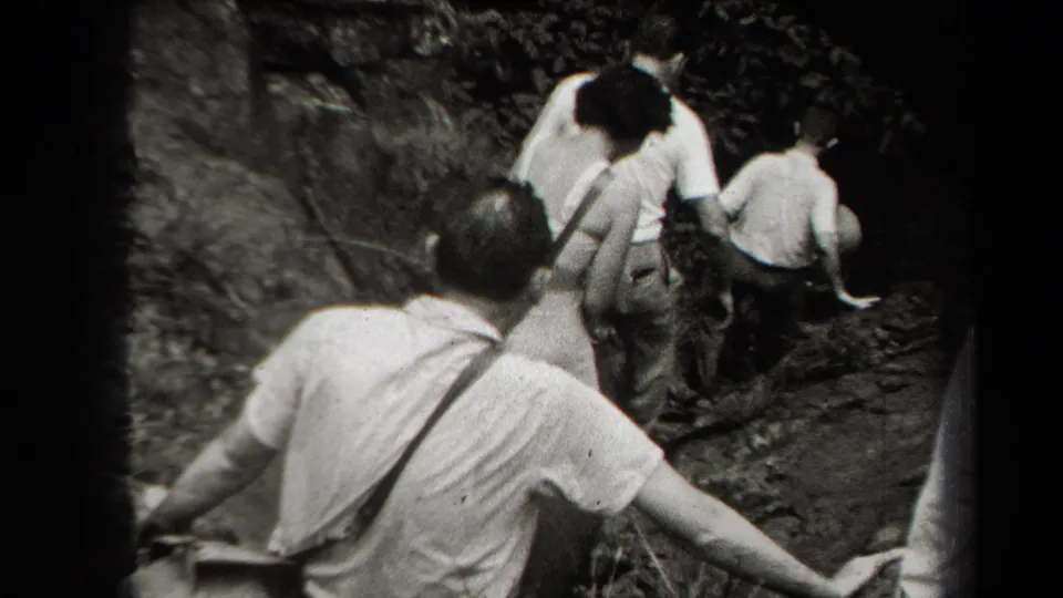 Adults move farther into the rocky cave passage in black-and-white 8mm footage, with a shoulder bag and light clothing visible from behind.