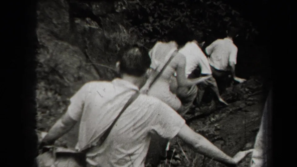 1938 New York home-movie frame showing men in light shirts moving up a steep rocky hillside through foliage.