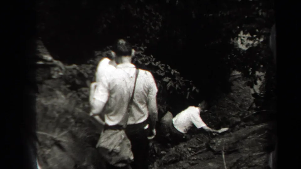 1938 New York footage frame with hikers navigating uneven rocks and brush, one figure crouched near the trail.