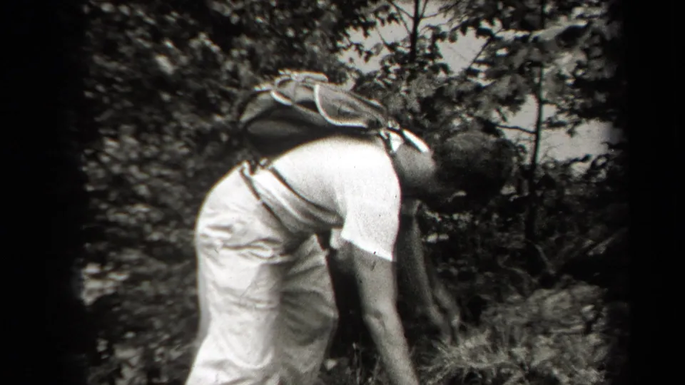 1938 New York frame showing a man with a small pack bending over on a rocky, wooded hillside.