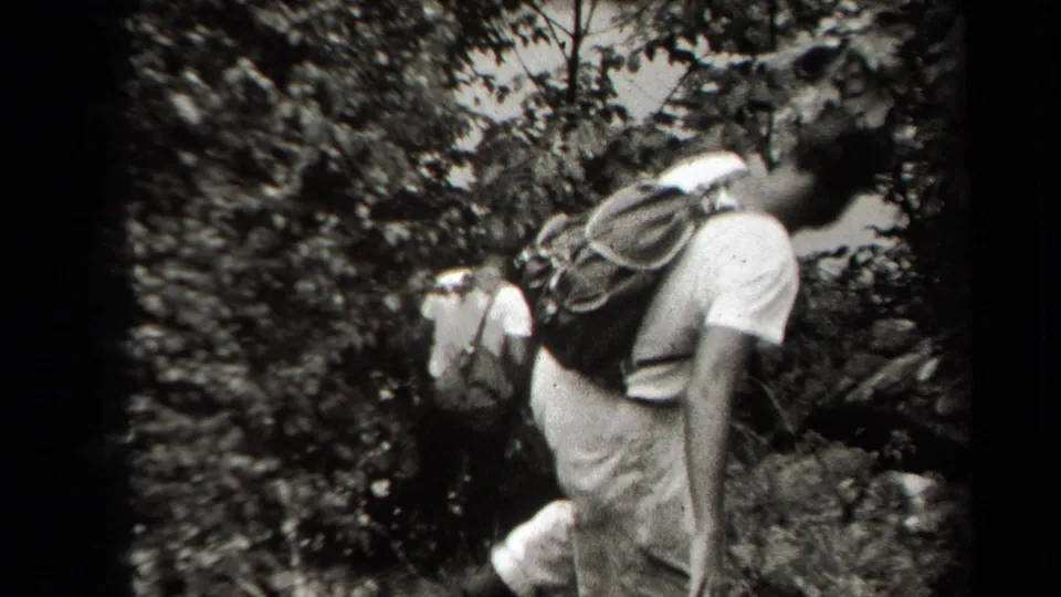 1938 New York home-movie frame with a backpacked hiker holding a stick on a rocky hillside.