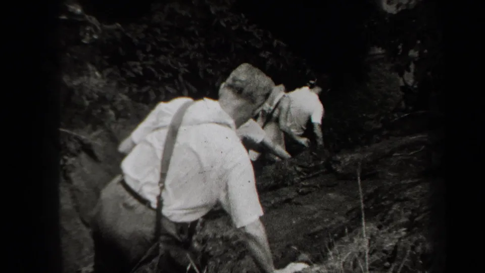 1938 New York 8mm still showing a man in suspenders descending behind others on uneven wooded terrain.