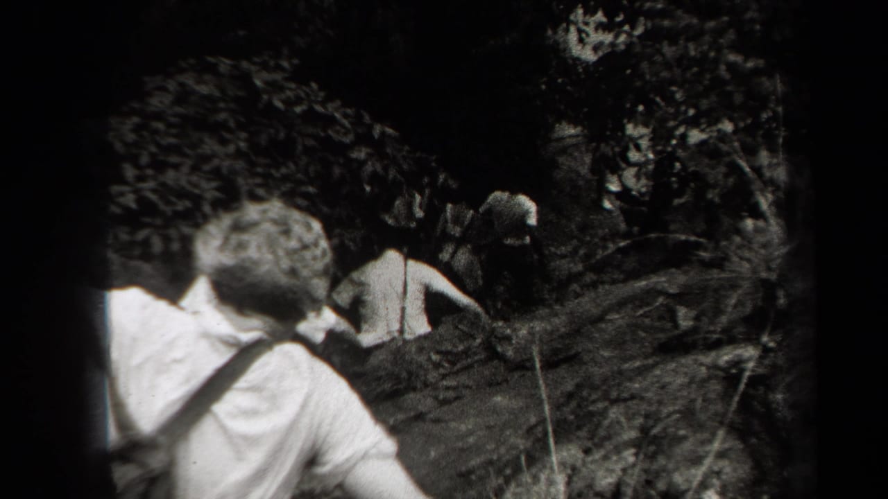 Hikers move through a dark rocky woodland trail, seen from behind in black-and-white footage.