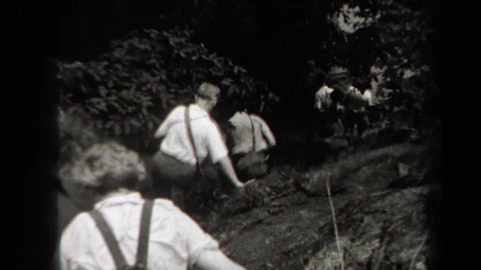 Black-and-white 8mm frame of friends hiking uphill through rocky woods in New York, with figures mostly seen from behind.