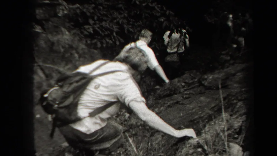 Black-and-white 1938 New York frame of a hiker using a hand for balance while following friends through rocky forest terrain.
