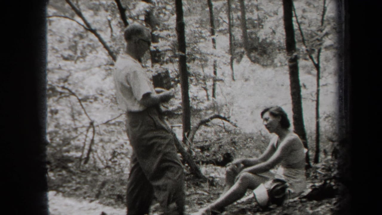 Man standing with a camera near a seated woman in a wooded area.