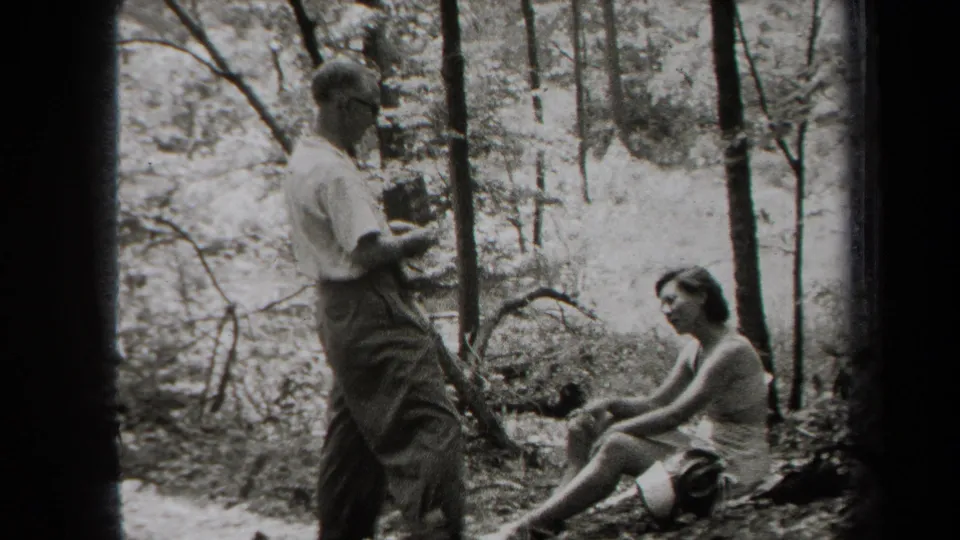 In supported 1938 New York home-movie footage, a man stands with a small camera while a woman sits on the forest floor nearby.