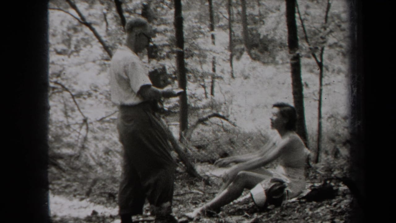 Man holding a camera faces a seated woman among trees.