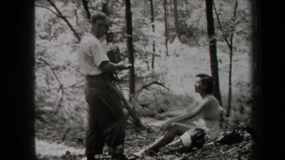 A man in a light shirt holds a small camera while a woman sits on the ground in a wooded New York setting, based on supplied metadata.