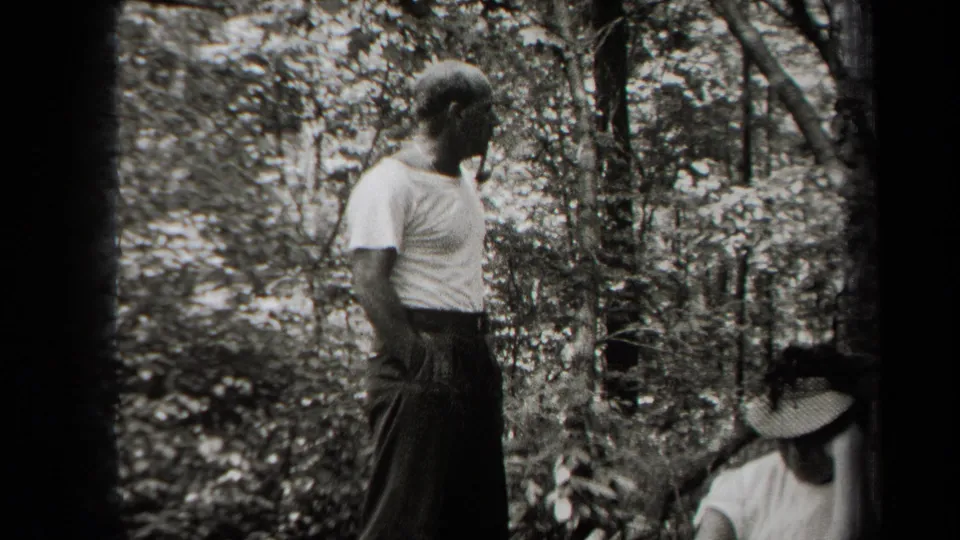 The man looks across a wooded area as the seated woman, partly shaded by a brimmed hat, appears near a bag or belongings.