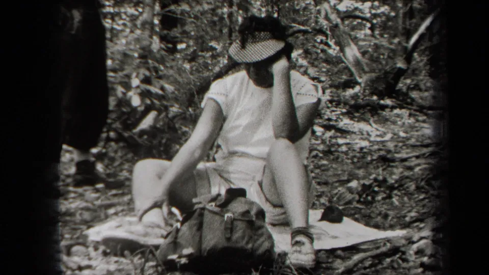 A seated woman in light clothing adjusts a brimmed hat beside a handbag on the forest floor in the 1938 home-movie scene.