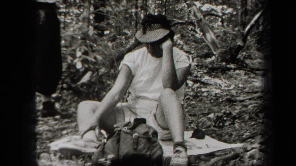In 1938 New York home-movie footage, a woman sits on a blanket in a wooded area and adjusts or holds her sun hat.