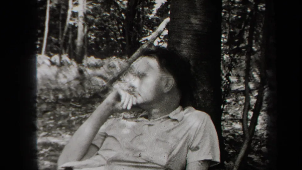 A man wearing glasses and a collared shirt rests near a tree, looking to the side in wooded 1938 New York footage.
