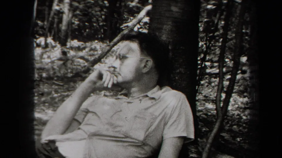 In a wooded New York setting, a man in glasses and a light collared shirt rests against a tree and looks away from the camera.