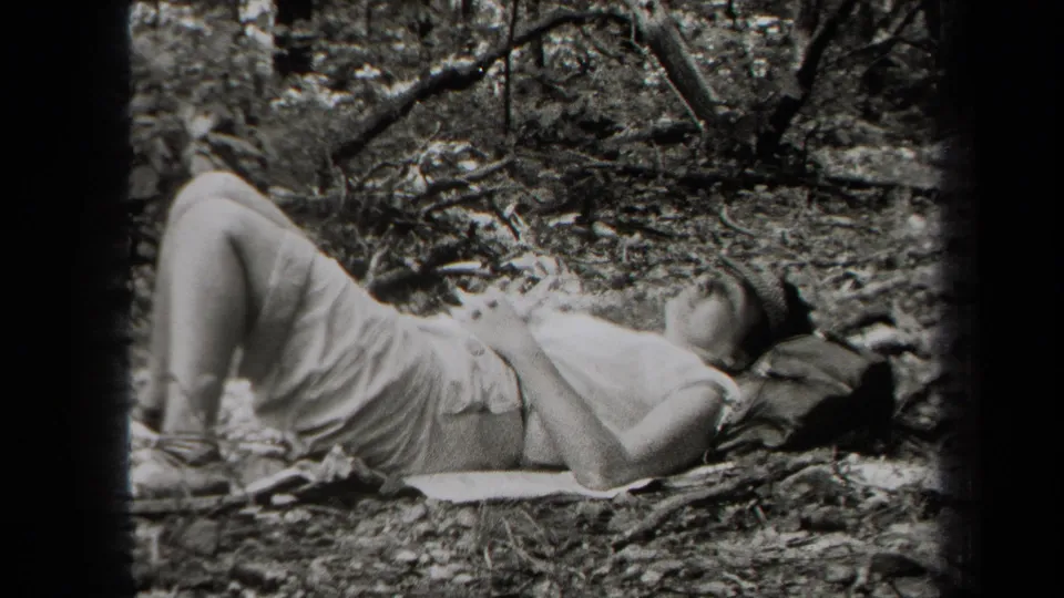 1938 New York home-movie frame of a person reclining on a blanket on the forest floor, surrounded by leaves and branches.