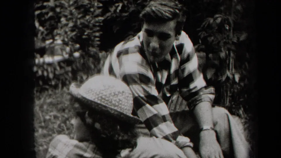 1938 New York home-movie frame of two people seated outdoors, with a striped shirt and brimmed hat visible in a shaded wooded area.