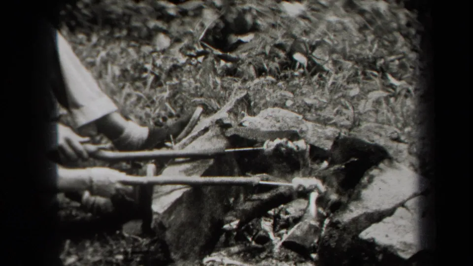 Black-and-white 8mm frame from 1938 New York showing hands using two sticks to roast food over a small fire outdoors.