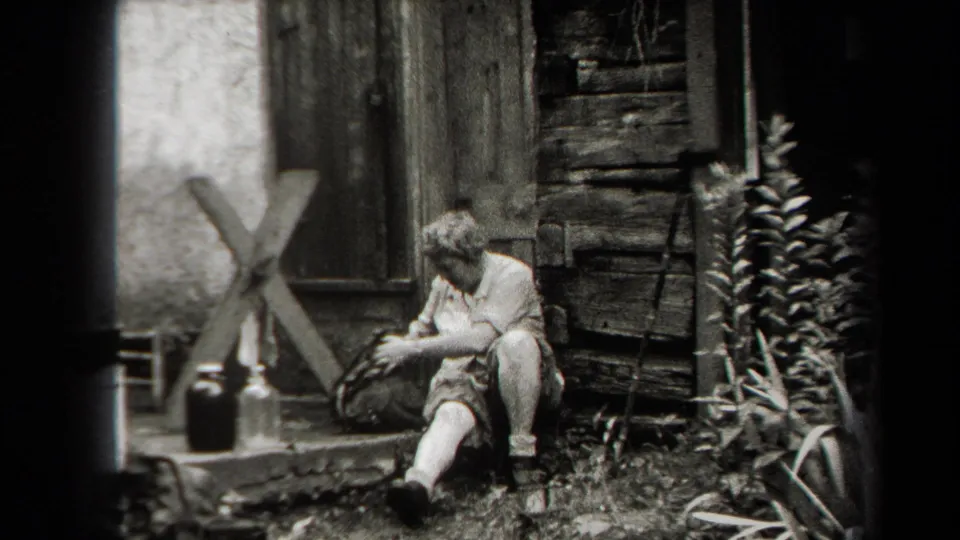 A woman sits outdoors beside a rustic wooden building with containers nearby in metadata-supported 1938 New York footage.