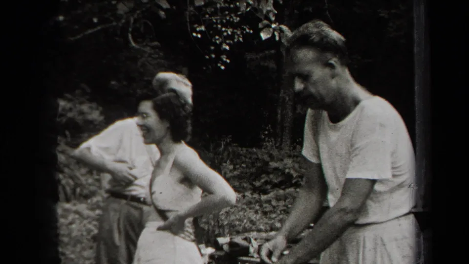 A small group stands in a wooded outdoor setting while a man tends food on sticks in metadata-supported 1938 New York footage.