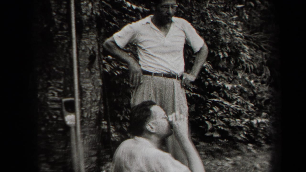 In supported 1938 New York home-movie footage, two adult men appear in a shaded backyard, one standing while another sits below him.