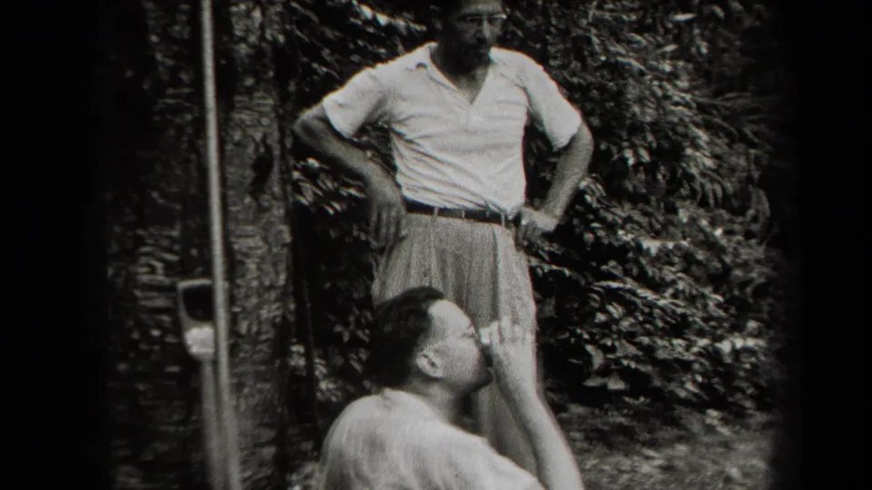 In supported 1938 New York home-movie footage, two adult men appear in a shaded backyard, one standing while another sits below him.