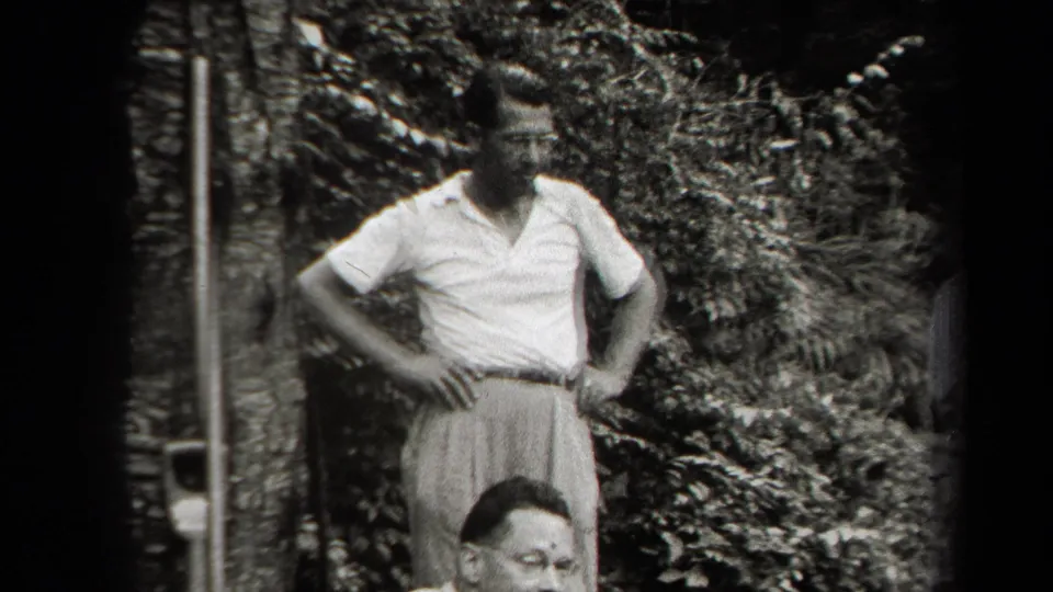 A man in a light shirt and pleated trousers stands in a leafy backyard, with another adult partly framed at the bottom.