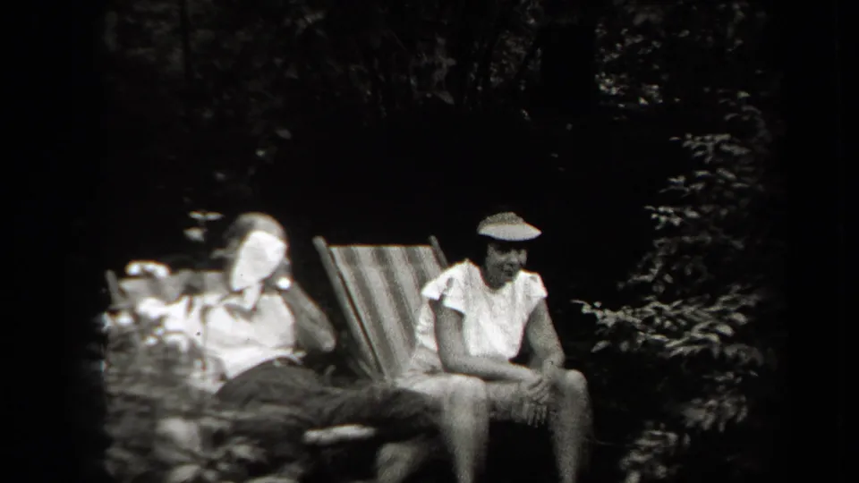 Two adults relax in outdoor chairs in the shaded yard, including a person in a cap seated beside a striped chair.