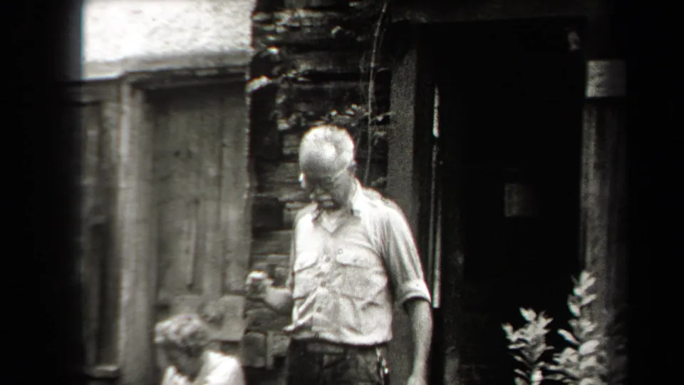 A man stands near a grill or outdoor stove by a rustic structure in the backyard, looking down at something in his hand.
