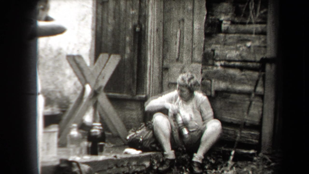 Woman seated on a step beside bottles and a bag near a weathered wooden structure.