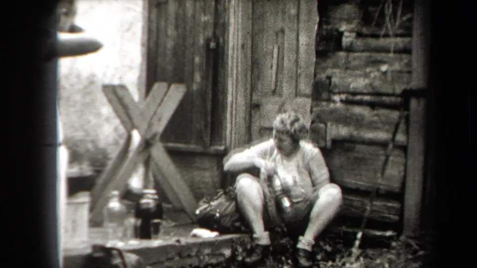 1938 New York home-movie frame showing a woman seated outdoors beside bottles, a bag, and rough wooden siding.