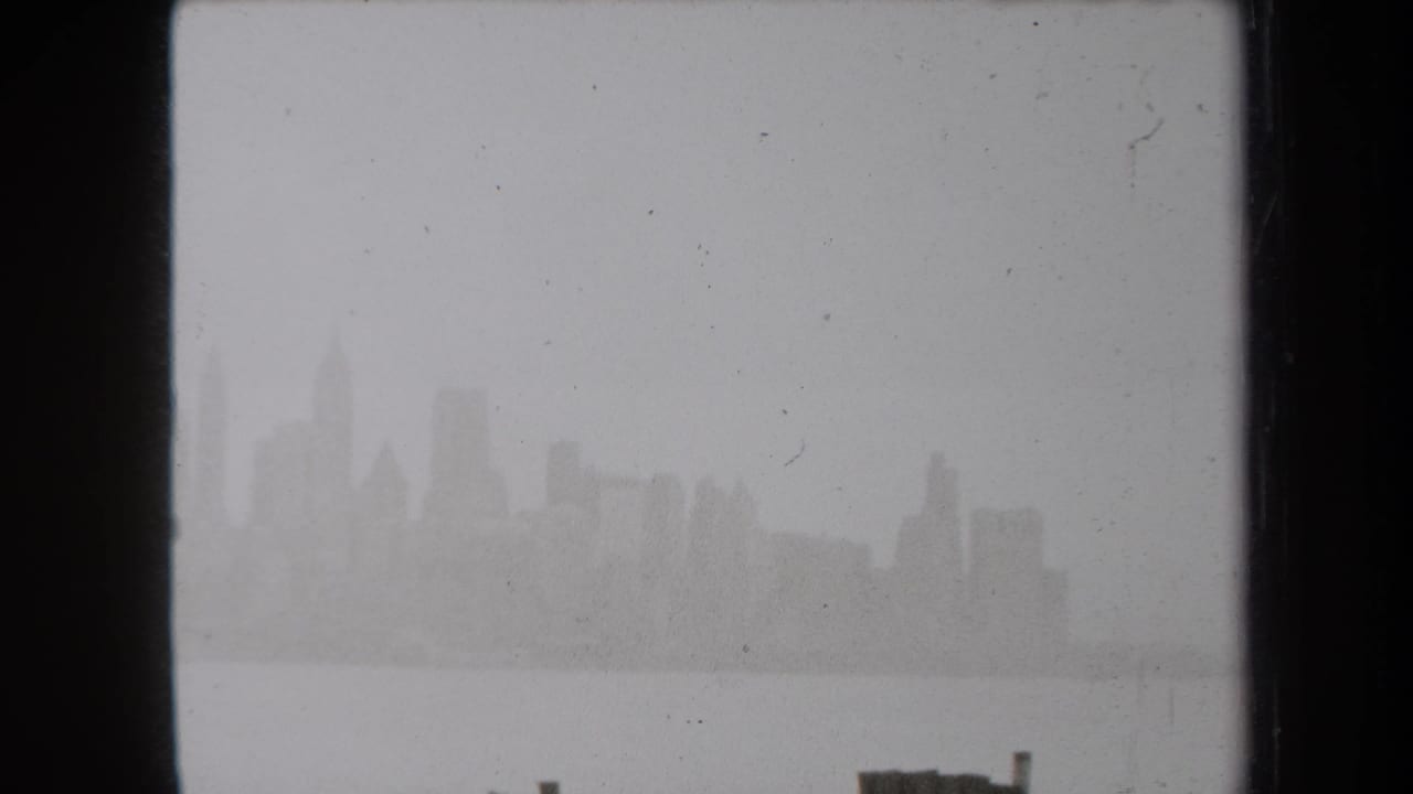 Fog-softened New York skyline across harbor water in 1938 8mm home-movie footage, with dark frame edges and dust specks.