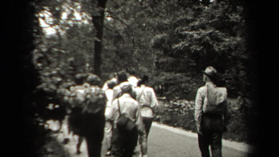 The handheld camera follows several adults from behind as they walk along a park path bordered by dense greenery.