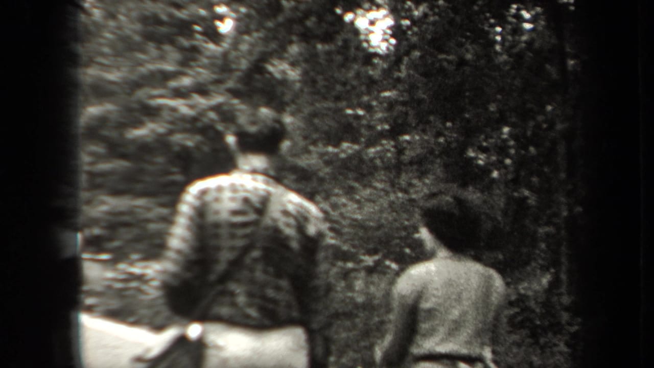 In 1938 New York home-movie footage, two adults walk away from the camera through a wooded area, with one backpack visible.