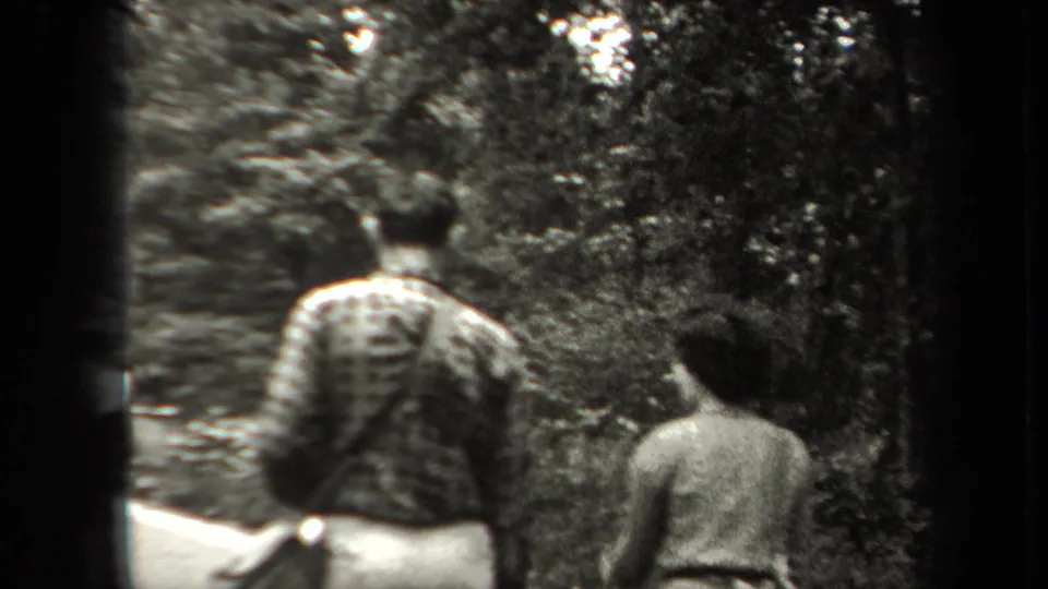 In 1938 New York home-movie footage, two adults walk away from the camera through a wooded area, with one backpack visible.