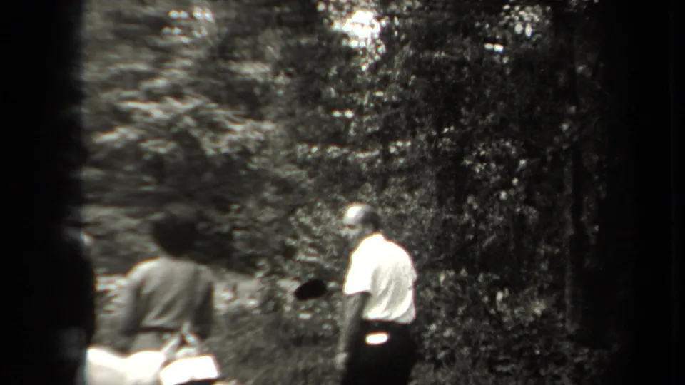 A small group pauses in the woods, with a man in a light shirt at right and another person carrying a bag or pack.