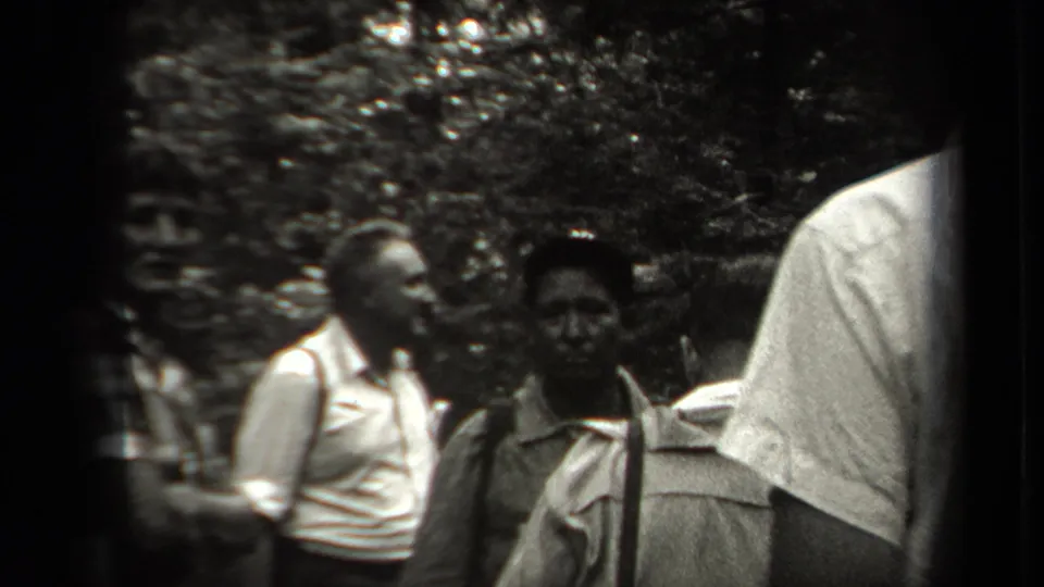 Adults in 1938 New York outdoor clothing gather in a wooded setting, including a man in a light shirt and a person facing the camera.