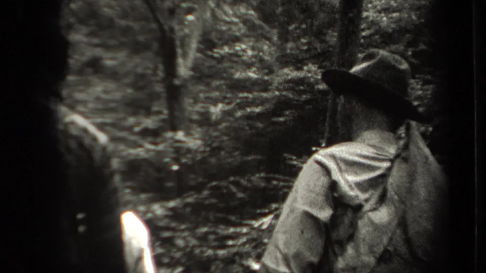 A person in outdoor clothing and a brimmed hat walks away through the New York woods in 1938 home-movie footage.