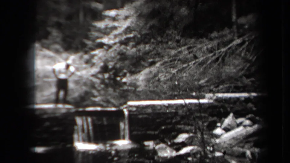 Vintage 8mm frame from New York in 1938 showing a distant man posed on a stone bridge amid trees and rocks.