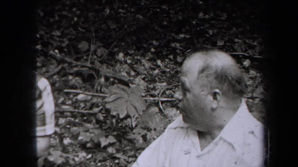 Black-and-white 1938 New York frame of a man in a light short-sleeved shirt beside a wooded hillside.