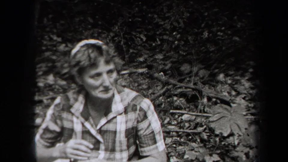 1938 New York home-movie frame showing a woman in a plaid collared dress resting near a leaf-covered trail.
