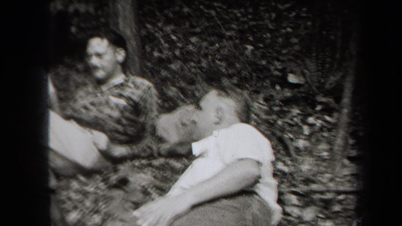 Adults seated on a wooded ground, with a man in a plaid shirt and another man in a light shirt visible.